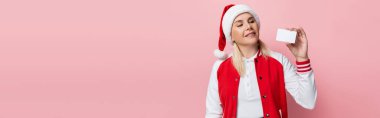 Positive woman in santa hat looking at empty driving license isolated on pink, banner 