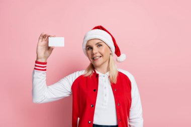 Blonde woman in red santa hat holding driving license with copy space isolated on pink 