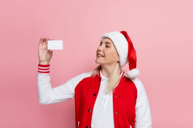 Smiling blonde woman in santa hat holding empty driving license isolated on pink 