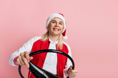 Smiling woman in santa hat holding steering wheel isolated on pink 
