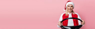 Positive woman in santa hat holding steering wheel isolated on pink, banner 