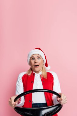 Scared woman in santa hat holding steering wheel isolated on pink 