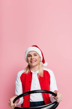 Cheerful woman in red santa hat holding steering wheel isolated on pink 