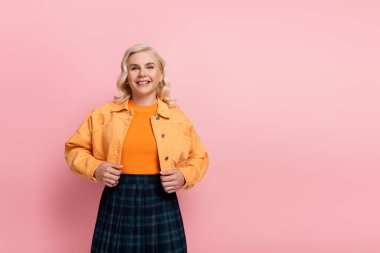 Smiling blonde woman in jacket looking at camera isolated on pink 