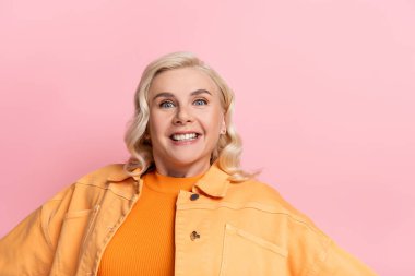 Cheerful woman in jacket looking at camera isolated on pink 