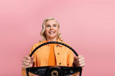 Smiling woman holding steering wheel and looking away isolated on pink 