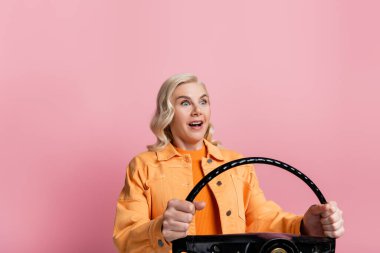 Shocked blonde driver holding steering wheel isolated on pink 