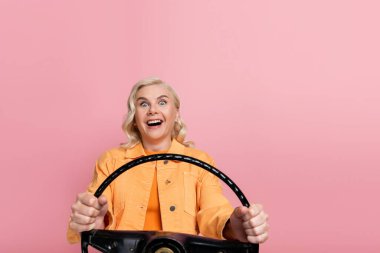 Amazed blonde driver holding steering wheel and looking at camera isolated on pink 