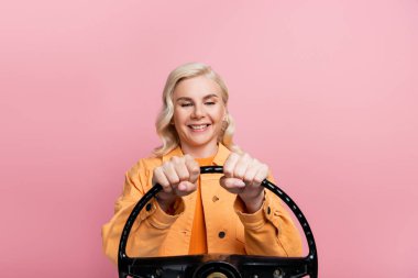 Positive blonde driver looking at steering wheel isolated on pink 