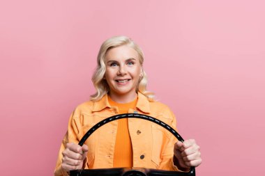 Happy blonde woman looking at camera while holding steering wheel isolated on pink 