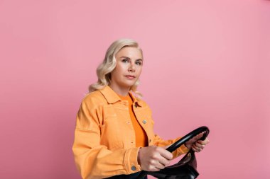 Blonde woman in jacket holding steering wheel isolated on pink 