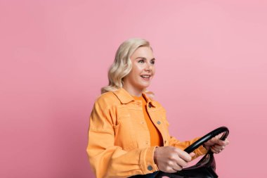 Blonde driver smiling and holding steering wheel isolated on pink 