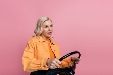 Blonde driver in orange jacket holding steering wheel isolated on pink 