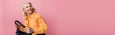 Smiling blonde driver in jacket holding steering wheel ad looking away isolated on pink, banner  