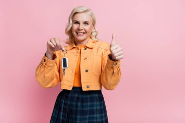 Smiling blonde driver holding car key and showing like gesture isolated on pink 
