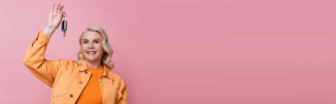 Cheerful blonde driver in jacket holding car key isolated on pink, banner 