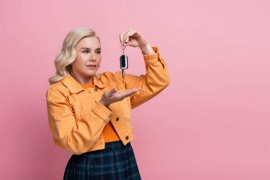 Blonde woman in jacket pointing at car key isolated on pink 