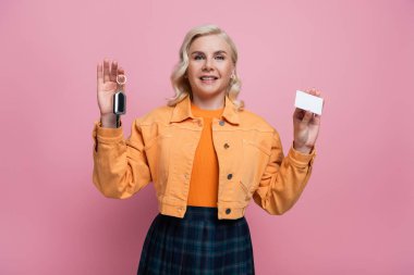 Smiling woman holding car key and driving license isolated on pink 