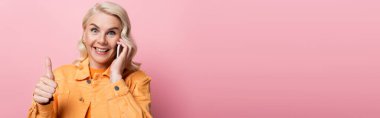 Positive woman showing thumb up and talking on smartphone isolated on pink, banner 