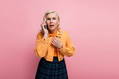 Scared blonde woman looking at camera while talking on smartphone isolated on pink 