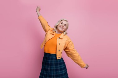 Positive woman in orange jacket stretching isolated on pink 