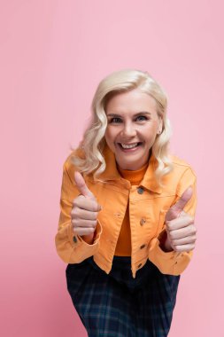 Cheerful blonde woman showing thumbs up at camera isolated on pink 
