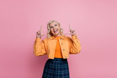 Positive woman in orange jacket pointing with fingers and looking up isolated on pink 