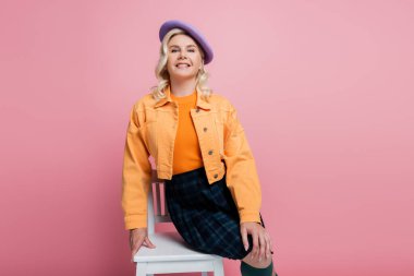 Happy woman in jacket and beret posing on chair isolated on pink 