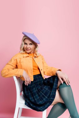Trendy young woman in beret sitting on chair on pink background