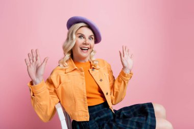 Astonished blonde woman in beret sitting on chair isolated on pink 