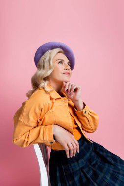 Dreamy woman in beret sitting on chair isolated on pink 