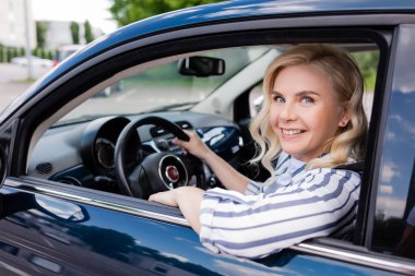 Cheerful blonde woman looking at camera while driving auto during course 