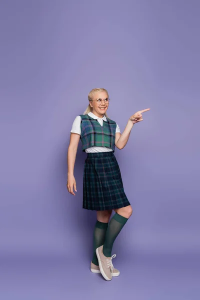 Full length of smiling student in uniform and eyeglasses pointing with finger on purple background