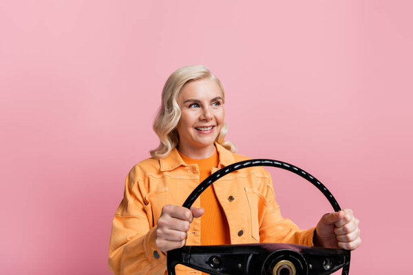 Blonde woman holding steering wheel and looking away isolated on pink 