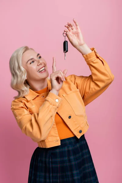 Positive blonde woman pointing at car key isolated on pink