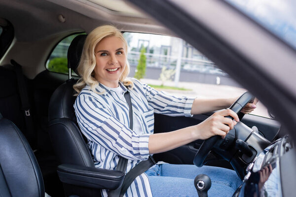 Cheerful blonde driver holding steering wheel and looking at camera in auto 