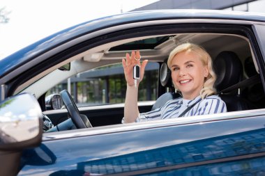 Positive blonde woman holding key while sitting in car 