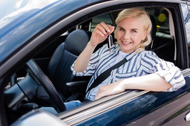 Cheerful blonde driver holding key while sitting in auto 