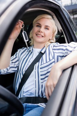 Positive blonde woman holding key while sitting in car 