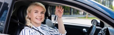 Positive blonde woman holding key and looking at camera from auto, banner 