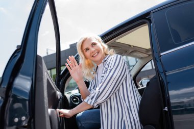 Smiling blonde woman waving hand while opening door of car outdoors 