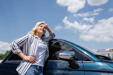 Low angle view of blonde woman closing eyes while standing near car outdoors 