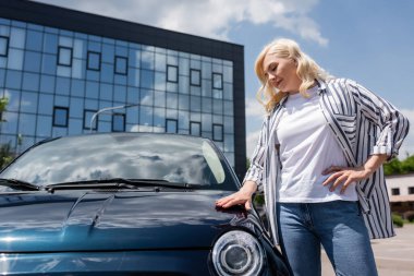 Blonde driver in shirt touching car on urban street 