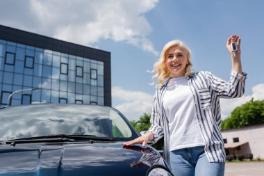 Smiling woman holding key near auto on urban street 