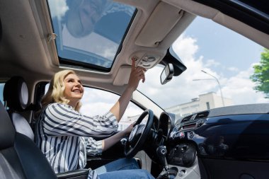 Cheerful blonde woman driving and looking at top of auto 