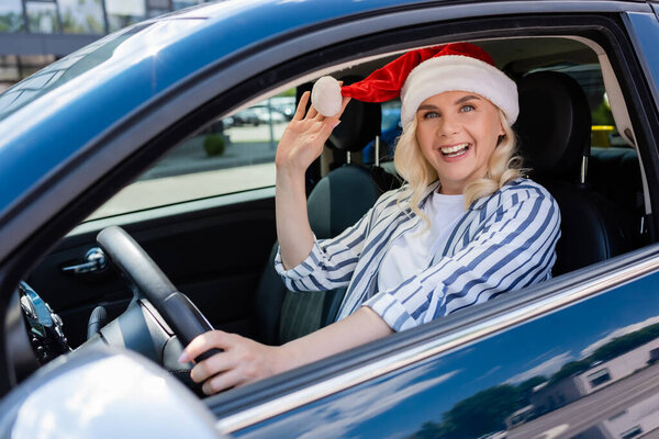 Positive blonde driver in santa hat looking at camera while driving car 