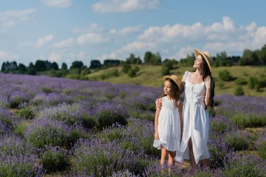 smiling woman in straw hat and white dress embracing daughter in lavender meadow