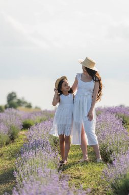 girl and mom in summer dresses and straw hats smiling at each other in blooming meadow