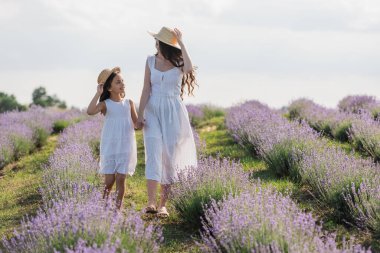 happy mom and daughter in dresses and straw hats holding hands and walking in meadow