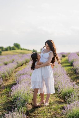 full length of brunette mom and daughter in white dresses embracing in meadow